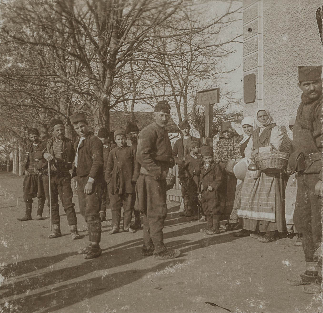 Bevolking in lokale klederdracht met op de achtergrond een bord met cyrillische tekst, 1912-1913. 